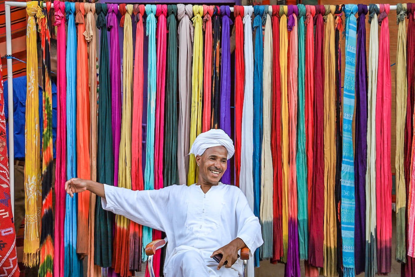 A Berber Amazigh man sits in front of his shop and colourful scarves and shawls hang behind him - an itinerary for 14 days in Morocco
